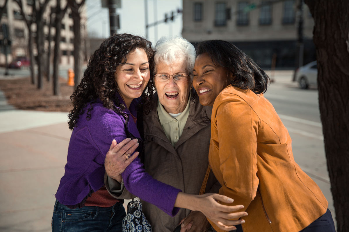 Senior woman hugged by two younger women on street corner